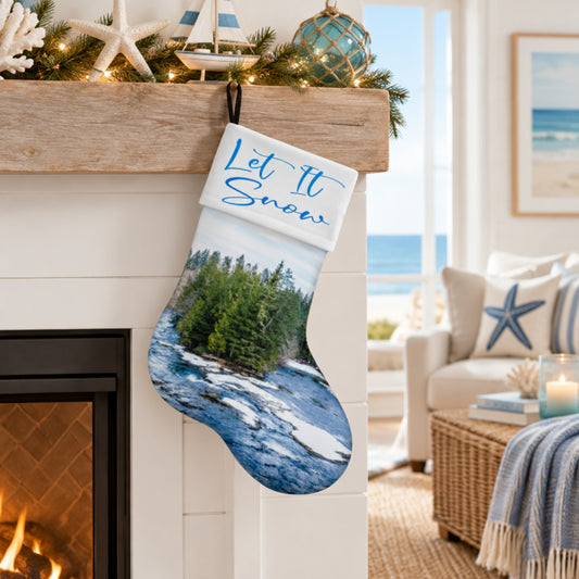 A Christmas stocking with a nautical landscape print and the text 'Let It Snow' hangs from a wooden mantel in a cozy living room with a fireplace, beach view, and coastal decor.