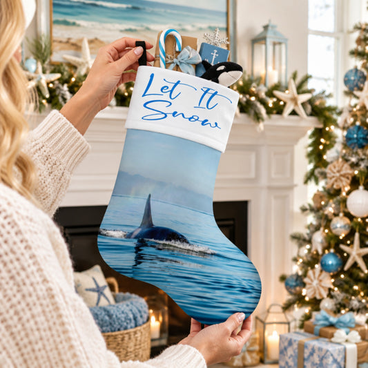 A person in a cream knit sweater holds up a Christmas stocking decorated with an orca whale print and the words 'Let It Snow' in blue script on a white cuff. The background shows a cozy, decorated holiday fireplace with a lit Christmas tree, gifts, and winter decorations.