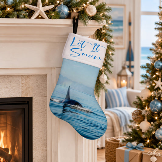 A Christmas stocking with a whale print and the embroidered text 'Let It Snow' on a white cuff, hanging in a decorated living room by a fireplace next to a lit Christmas tree.