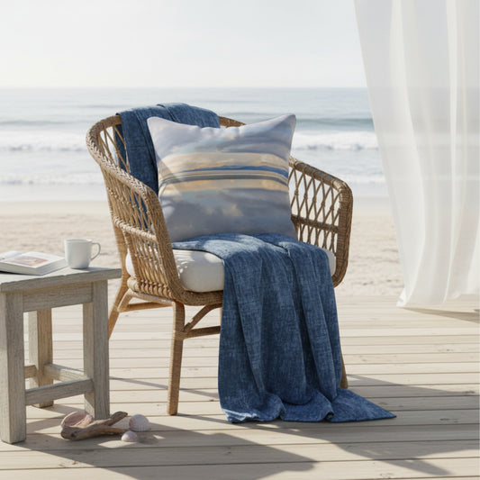 A woven outdoor chair on a beach deck with an ocean view, styled with a blue throw blanket, a striped blue-and-beige pillow, a small side table, and a white curtain.