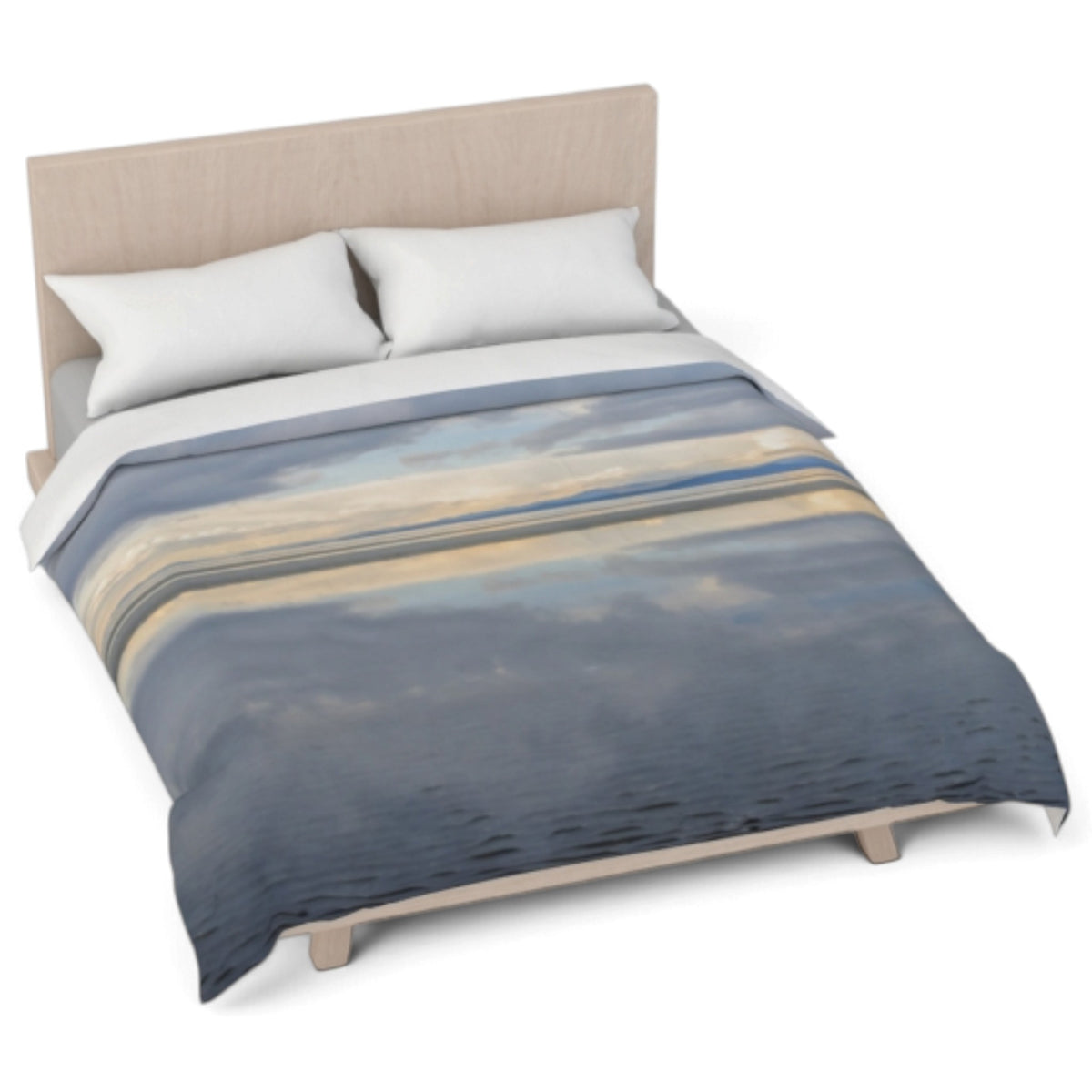 A comforter with a scenic landscape print of blue water, beige sand, and cloudy skies, styled on a bed with a light wood headboard and white pillows against a white background.