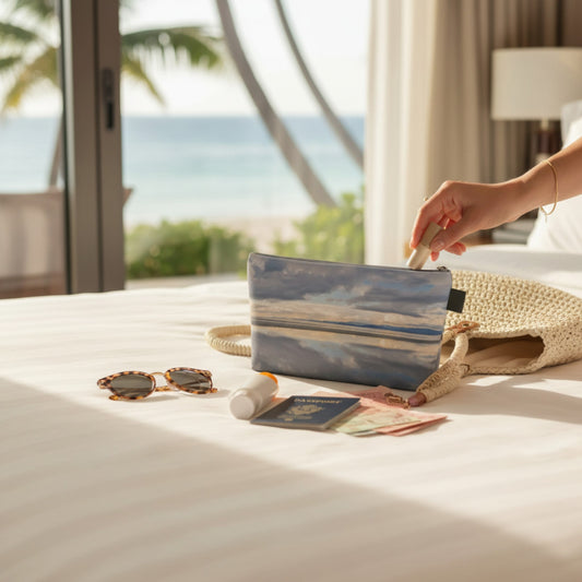Hand holding a travel pouch on a bed with sunglasses, passport, and other items.