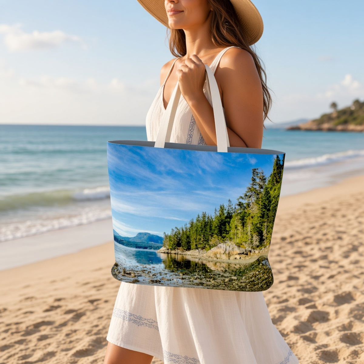 A woman on a sandy beach holds a large tote bag printed with a scenic landscape of a blue lake, green trees, and distant mountains. The woman wears a white dress and a straw hat, with the ocean and sky in the background.