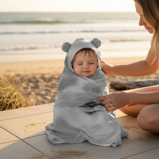 Baby wrapped in a gray hooded towel with bear ears, sitting on a wooden deck with a blurred beach background.