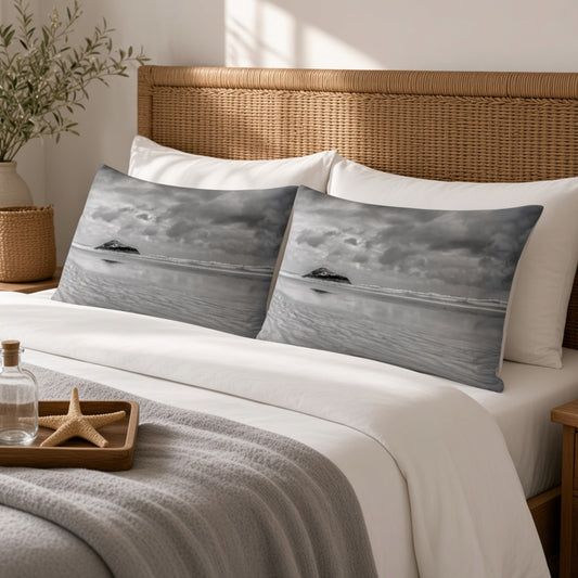 Two rectangular pillowcases with a grayscale ocean landscape print are styled on a bed with white linens, a wicker headboard, and a gray throw blanket in a bright bedroom setting.
