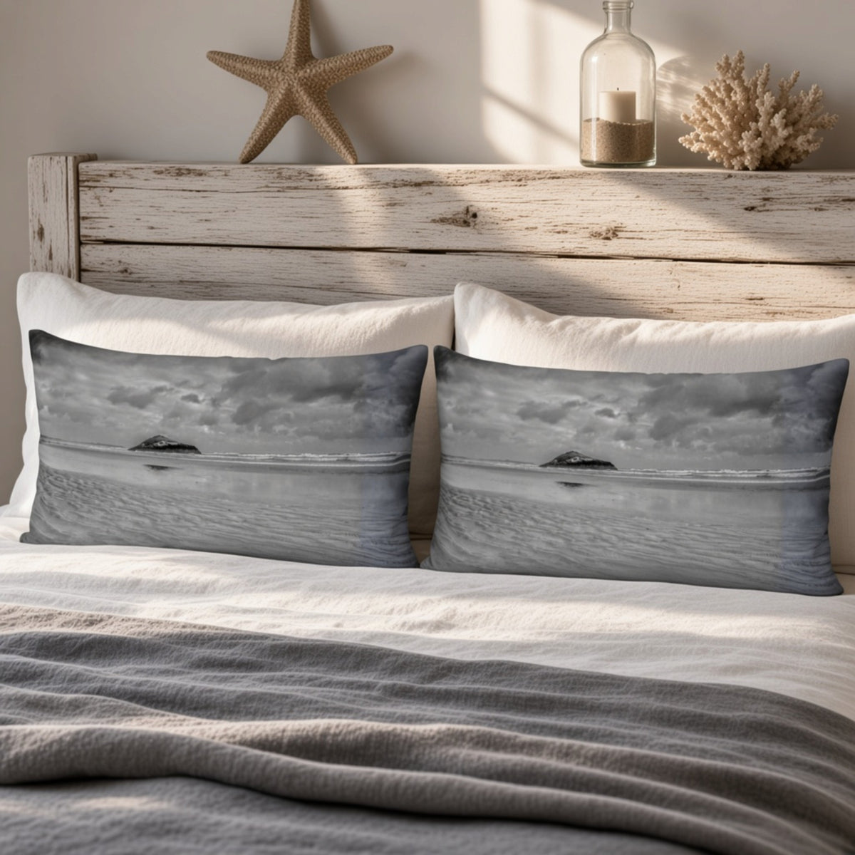 Two rectangular throw pillows with a grayscale ocean landscape print, styled on a bed with white pillows and a gray blanket in front of a rustic wooden headboard.