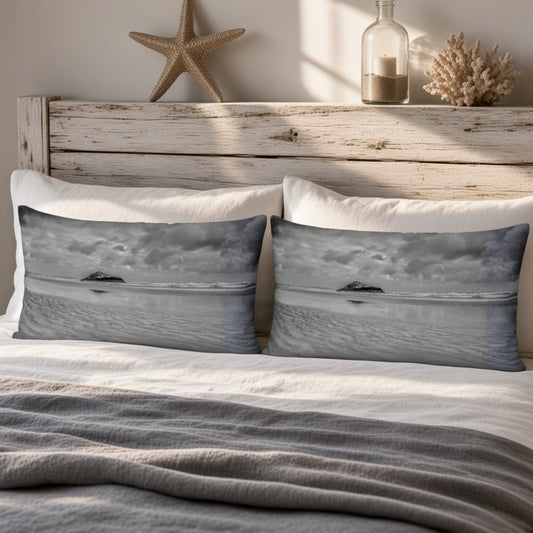 Two rectangular throw pillows with a grayscale ocean landscape print, styled on a bed with white pillows and a gray blanket in front of a rustic wooden headboard.