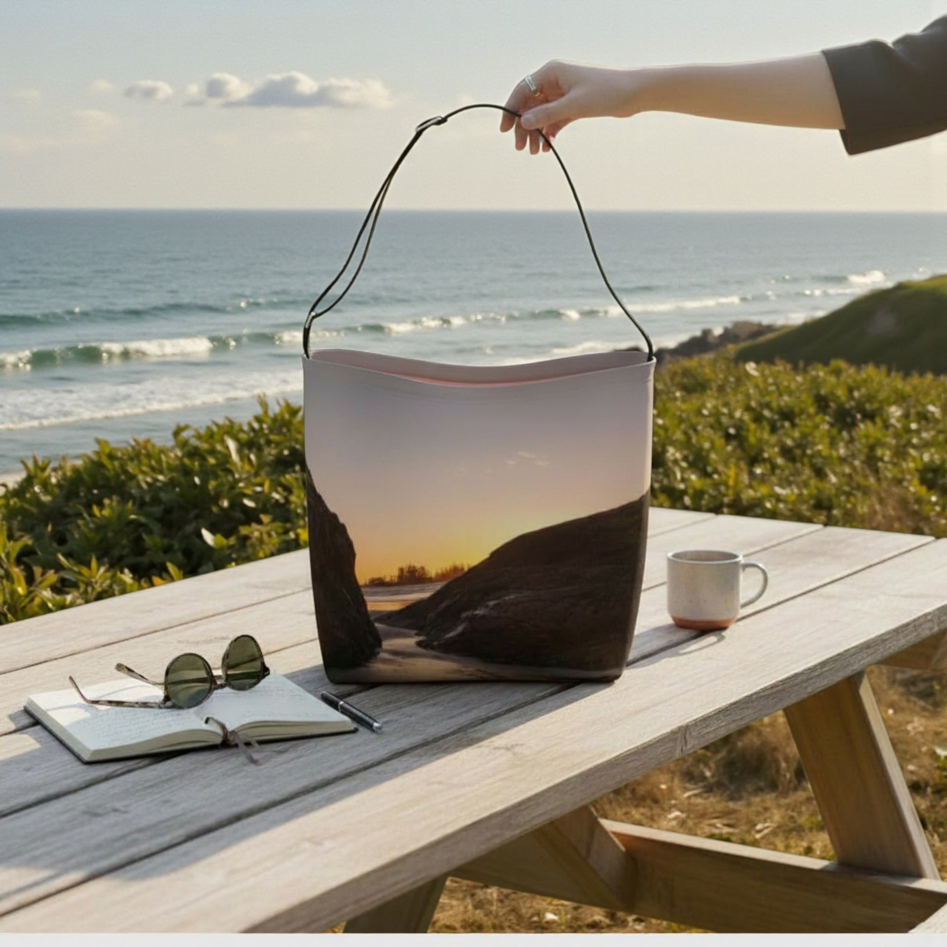 Person holding a bag with a sunset design on a wooden table by the ocean.