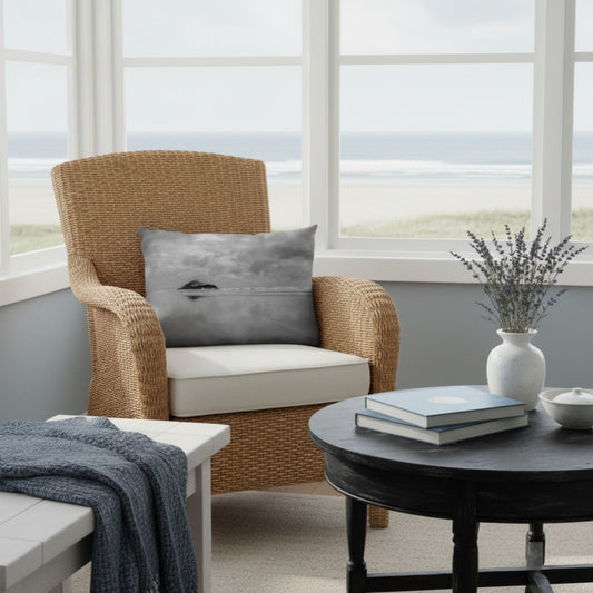 A gray throw pillow with a minimalist whale print rests on a woven wicker armchair in a bright, coastal living room setting.