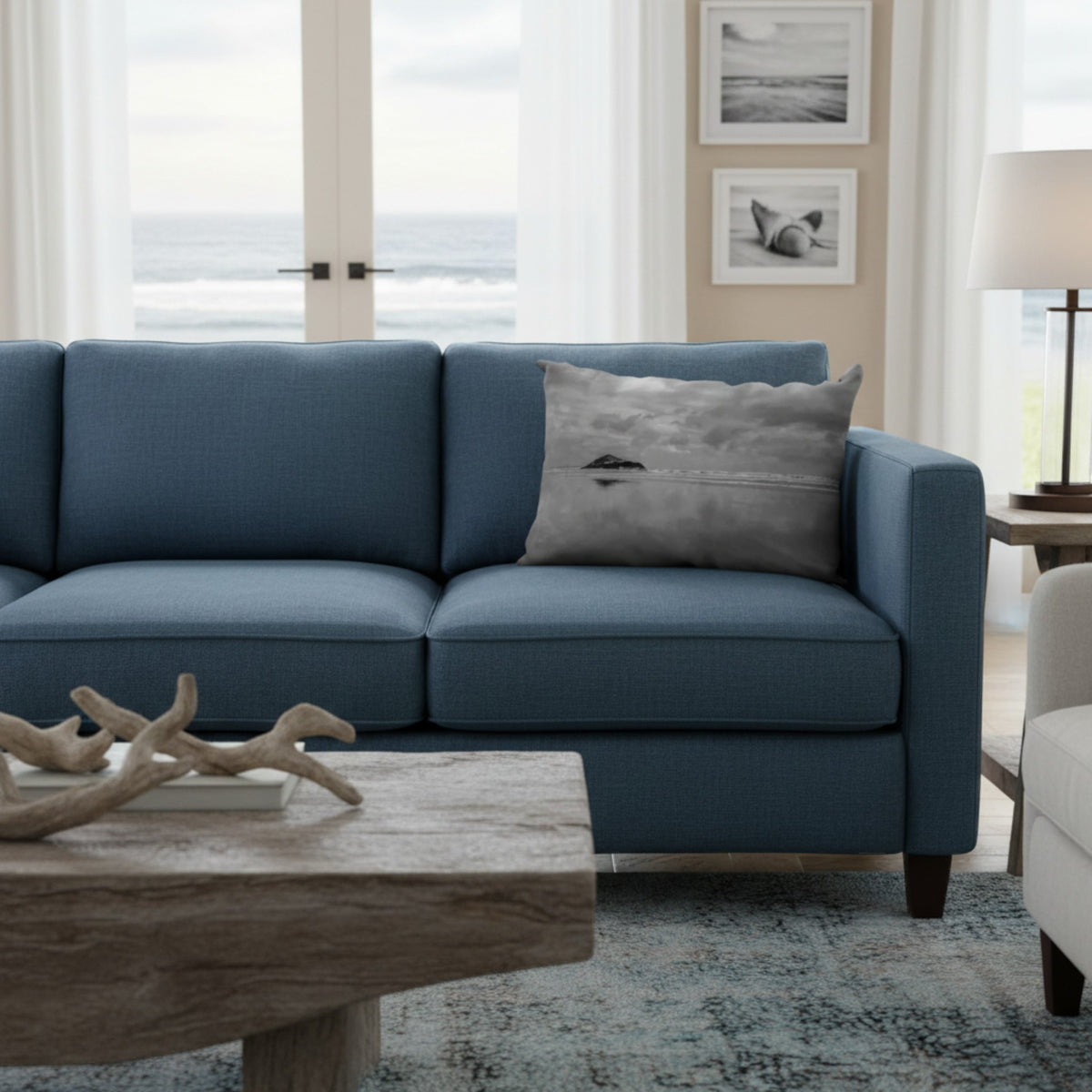 A blue fabric sofa in a bright, modern living room with an ocean view, featuring a gray throw pillow with a seashell design.