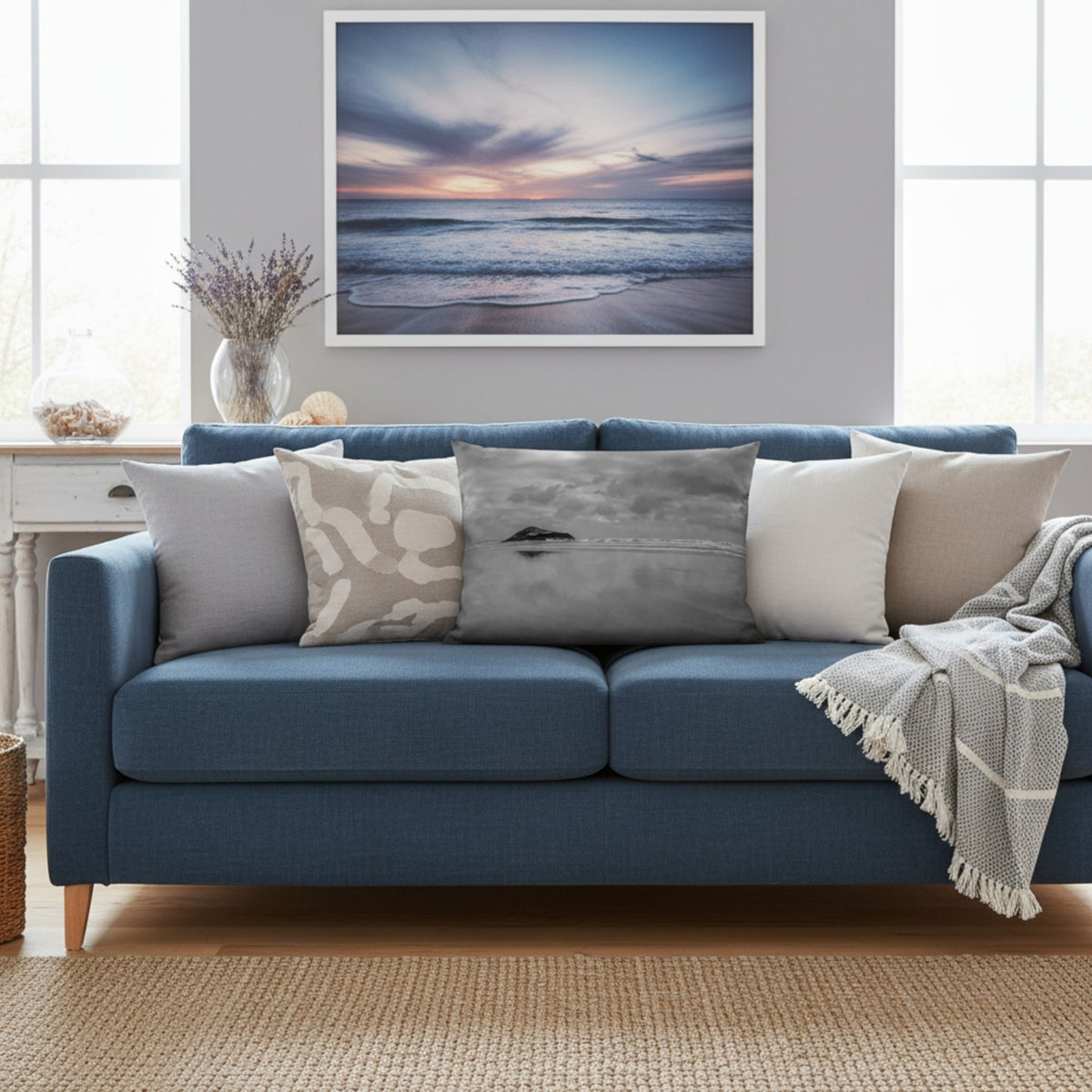 A framed square wall art print of a calm ocean sunset hangs above a blue sofa in a bright living room. The print shows soft waves at the beach under a pastel sky with pink, purple, and blue tones.