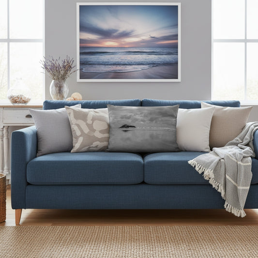 A framed square wall art print of a calm ocean sunset hangs above a blue sofa in a bright living room. The print shows soft waves at the beach under a pastel sky with pink, purple, and blue tones.