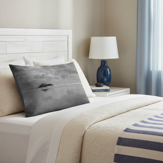 A square decorative throw pillow featuring a black and white landscape photograph of a small island on a calm sea, styled on a bed with cream bedding and a blue bedside lamp in a softly lit bedroom.