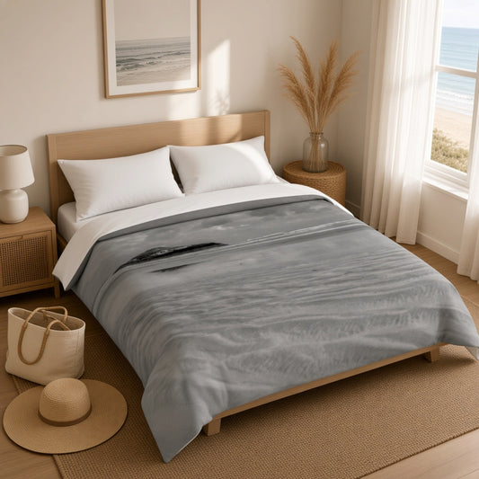 A gray and white watercolor-style blanket with a subtle wave pattern is styled on a light wood bed frame in a bright, modern bedroom with a window overlooking the ocean.