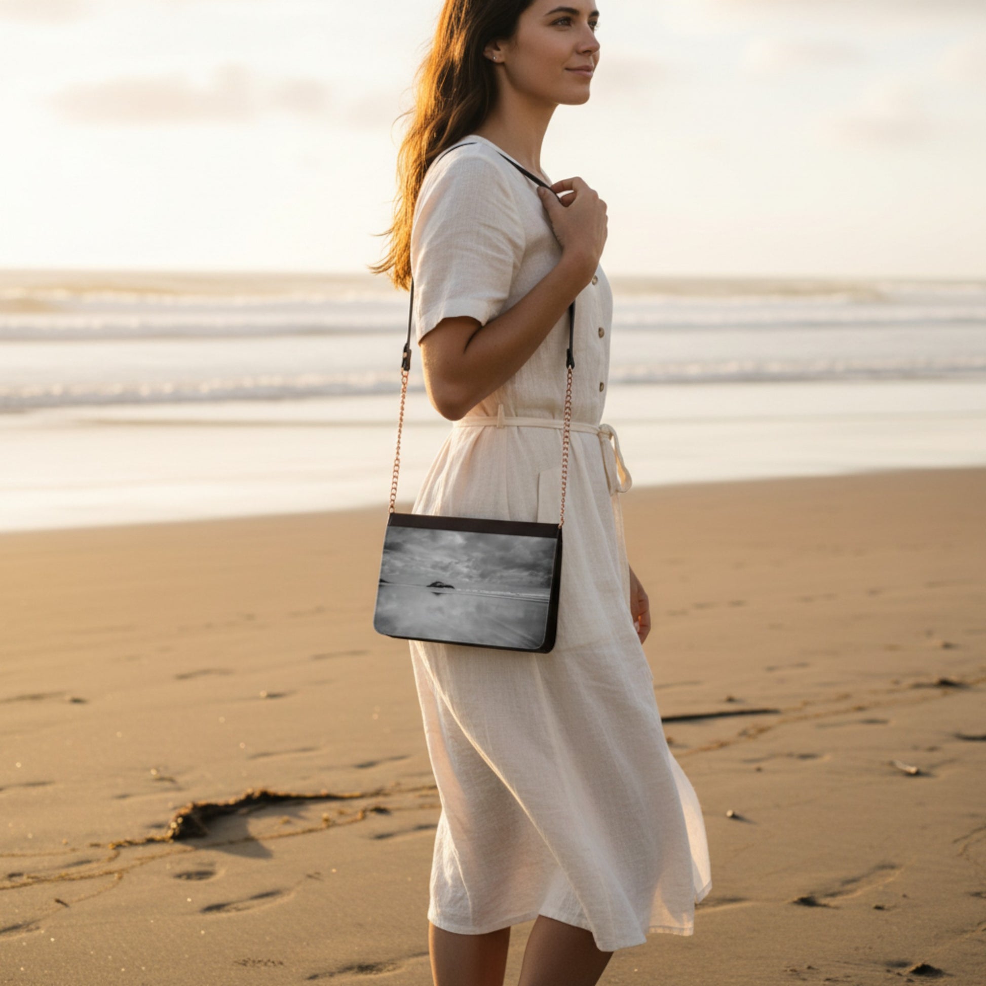 Woman in a white dress holding a black handbag with a scenic design on a beach.