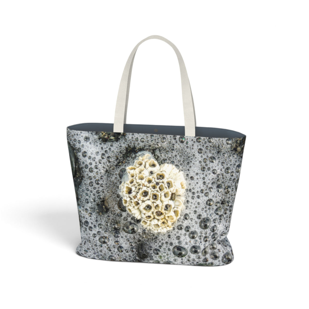 A tote bag featuring a photographic print of barnacles on wet sand, with a large, detailed sea urchin cluster printed on the front. The bag has light beige fabric handles and a dark navy blue interior lining, shown against a plain white background.