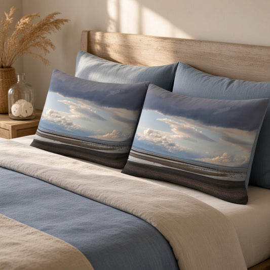 Two rectangular decorative pillows with a beach landscape print in blue, gray, and beige tones, styled on a bed with blue and beige bedding in a warm, sunlit bedroom.