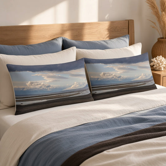 Two rectangular beach landscape throw pillows with a cloudy blue sky and dark shoreline print, styled on a bed with white and blue bedding in a warm, sunlit bedroom.