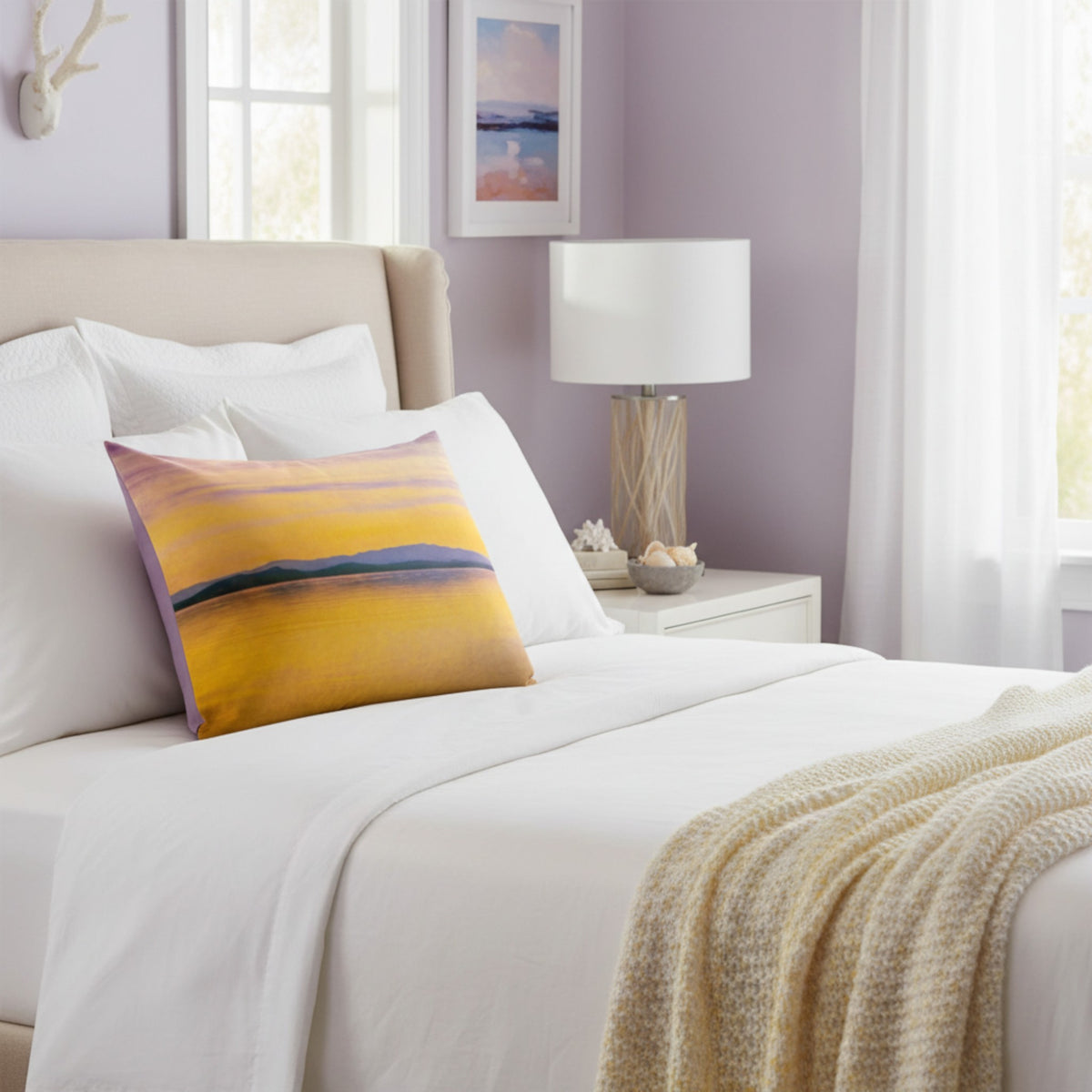 A square decorative throw pillow with an abstract landscape design in yellow, orange, purple, and blue, styled on a white bed in a bedroom with lavender walls and a beige headboard.