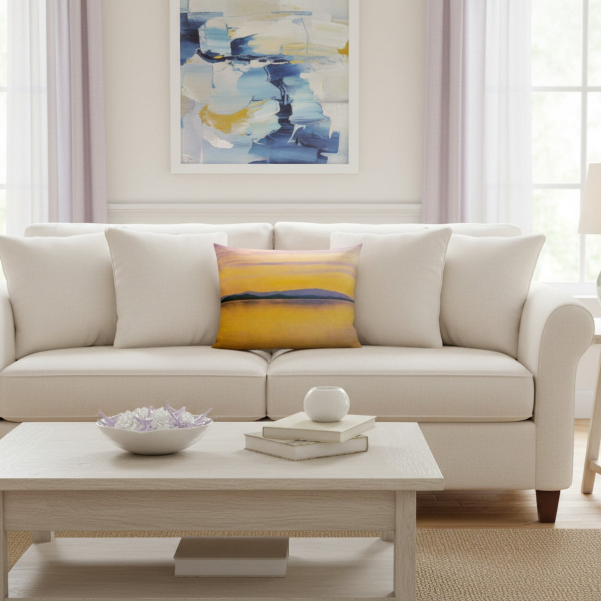 A cream-colored sofa in a bright, modern living room setting, styled with a square throw pillow featuring a yellow, pink, and blue abstract landscape design.