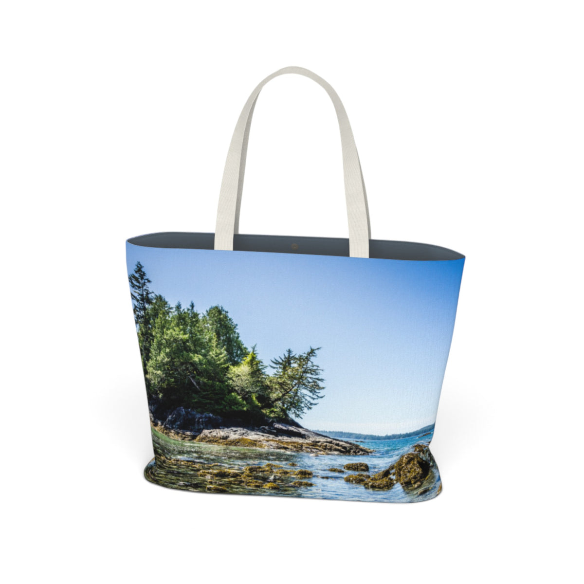 A tote bag with a printed coastal landscape design, featuring a rocky shoreline, green trees, blue sky, and ocean water. The bag has off-white shoulder straps and a dark interior, shown against a plain white background.