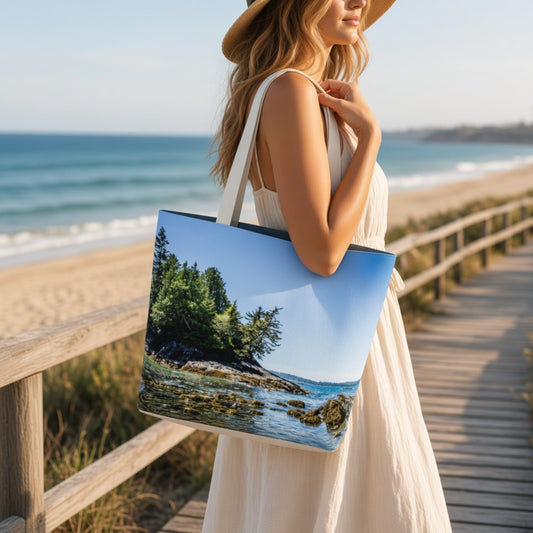 A woman on a wooden boardwalk by the beach carries a large tote bag printed with a scenic coastal landscape featuring green trees, rocks, and blue water under a clear sky.