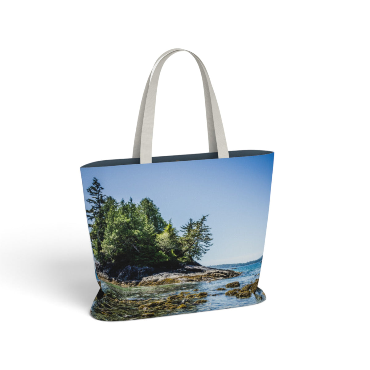 A tote bag featuring a printed scenic photograph of a rocky coastline with green trees, blue sky, and water, with light gray shoulder straps, shown on a white background.