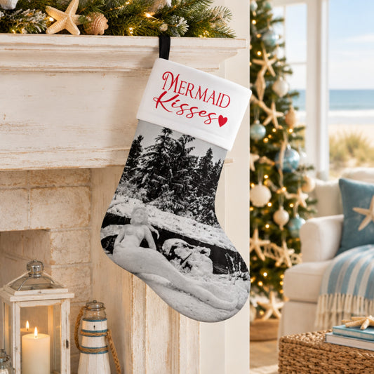 A personalized Christmas stocking hanging in a cozy living room setting next to a fireplace. The stocking features a white cuff with the words 'Mermaid Kisses' in red script and a black-and-white photo of a couple in a snowy landscape.