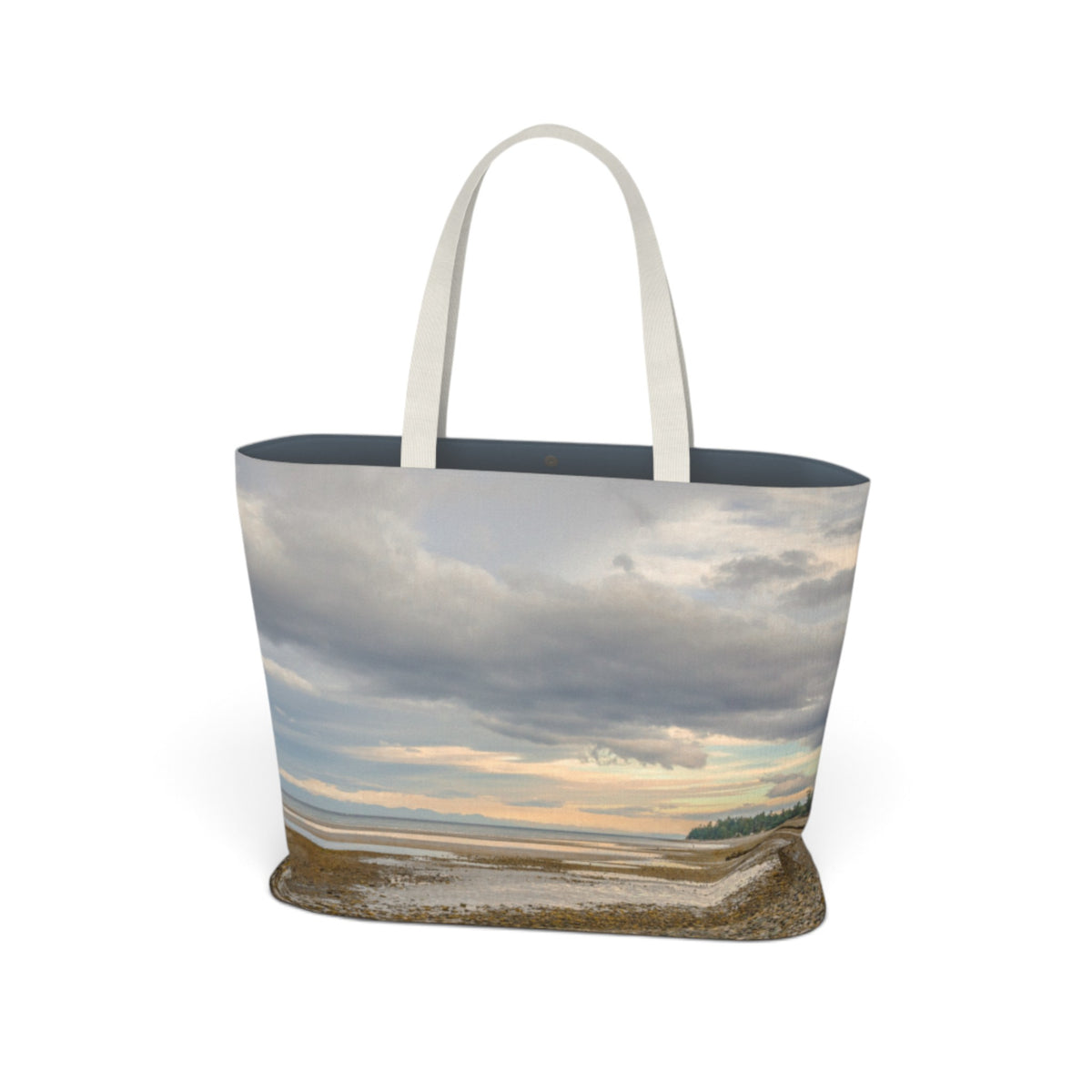 A tote bag featuring a photographic print of a coastal landscape with dramatic clouds, a pale sky, and wet sand, complete with two white shoulder straps, shown on a plain white background.