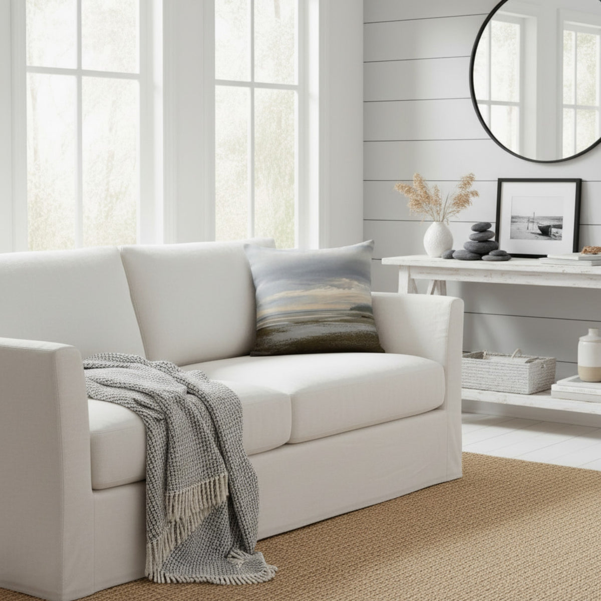 A white upholstered loveseat in a bright, modern living room, styled with a gray throw blanket and a square landscape pillow. The room features white shiplap walls, a white console table with decorative items, and a round black-framed mirror.