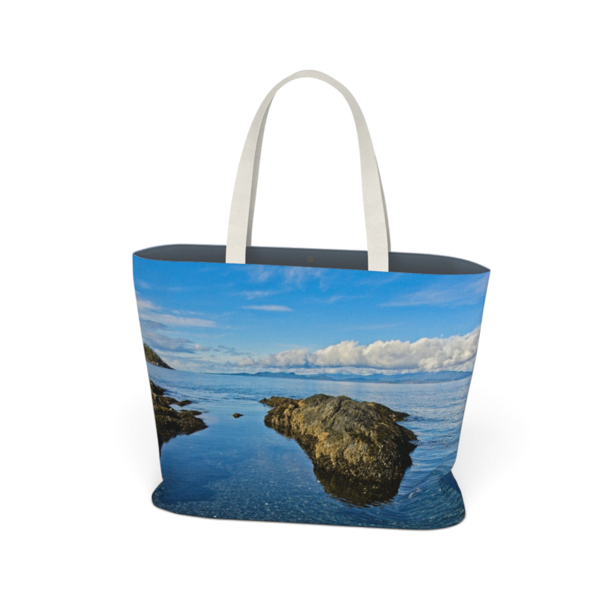 A tote bag featuring a scenic landscape print of a rocky coastline and blue water under a cloudy sky, with white shoulder straps, shown on a plain white background.