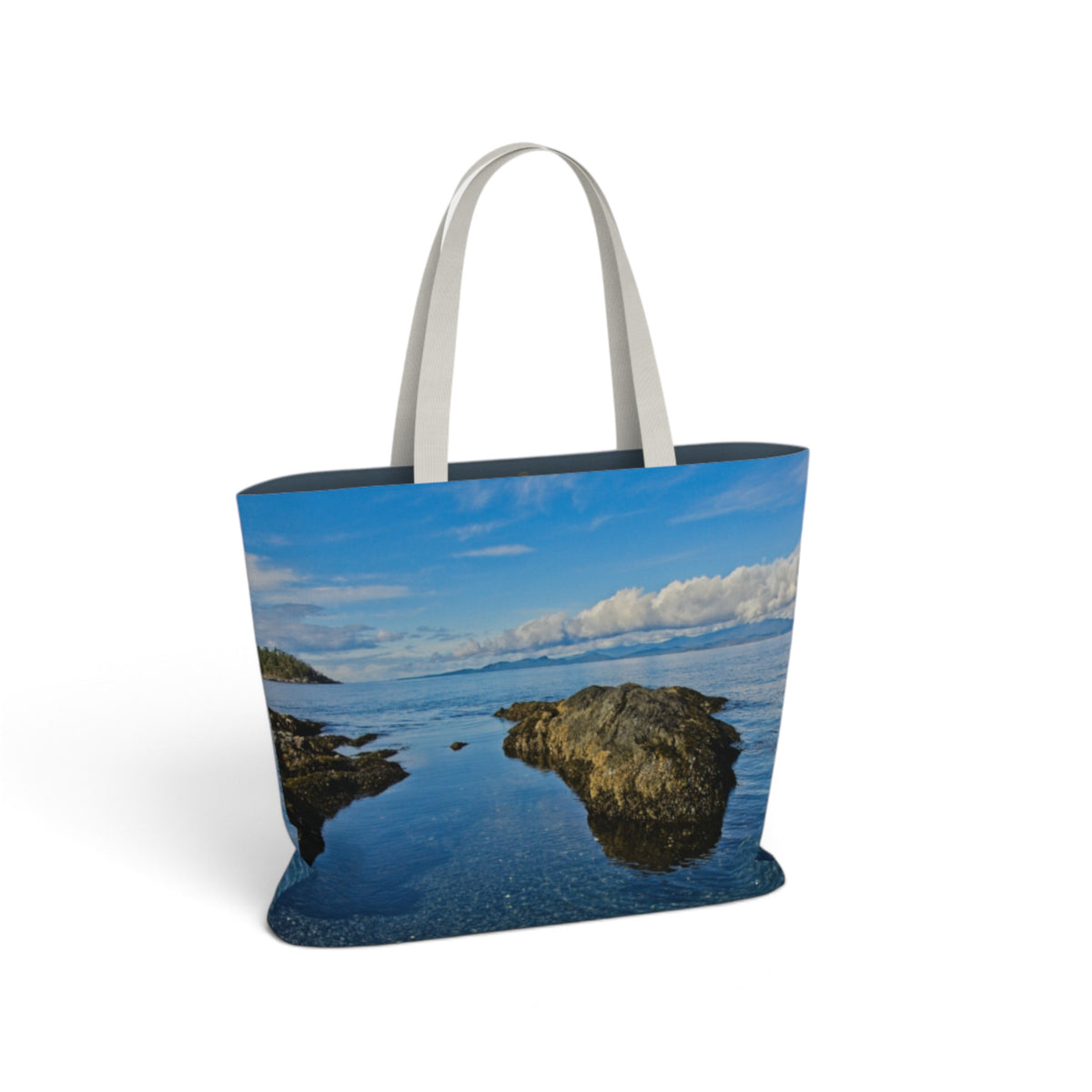 A tote bag featuring a scenic photograph of a rocky coastline and blue water, with light gray handles, shown on a plain white background.