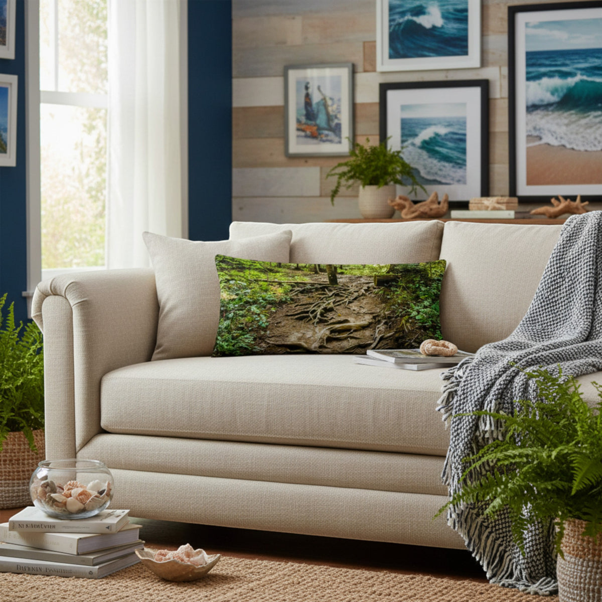 A beige upholstered loveseat in a styled living room, featuring a forest-patterned throw pillow, a gray knit throw blanket, and a woven rug. The background includes a wood-paneled wall with framed ocean art, potted plants, and a window.