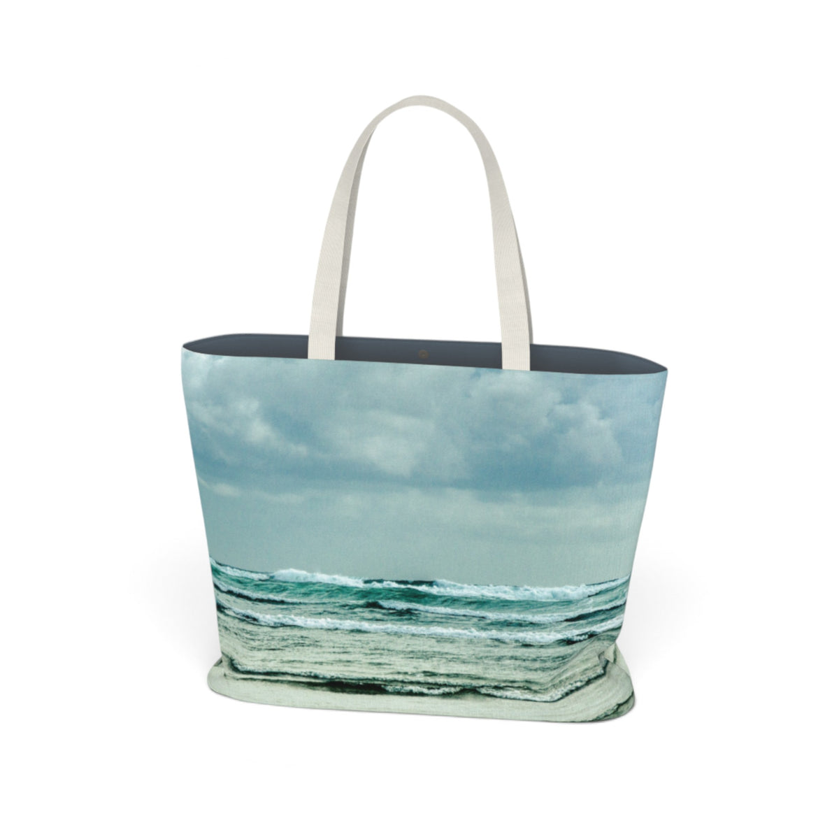 A tote bag featuring a printed beach and ocean scene with blue-green waves under a cloudy sky, finished with light beige shoulder straps, shown on a plain white background.