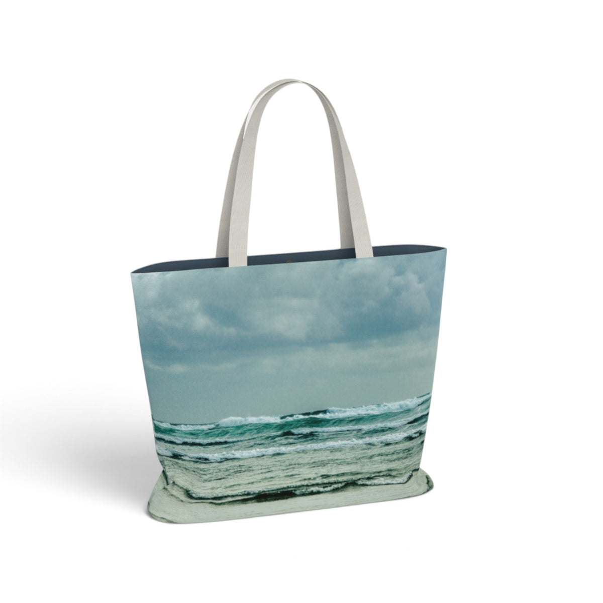 A tote bag featuring a printed beach and ocean scene with waves under a cloudy sky, with light beige shoulder straps, shown on a plain white background.