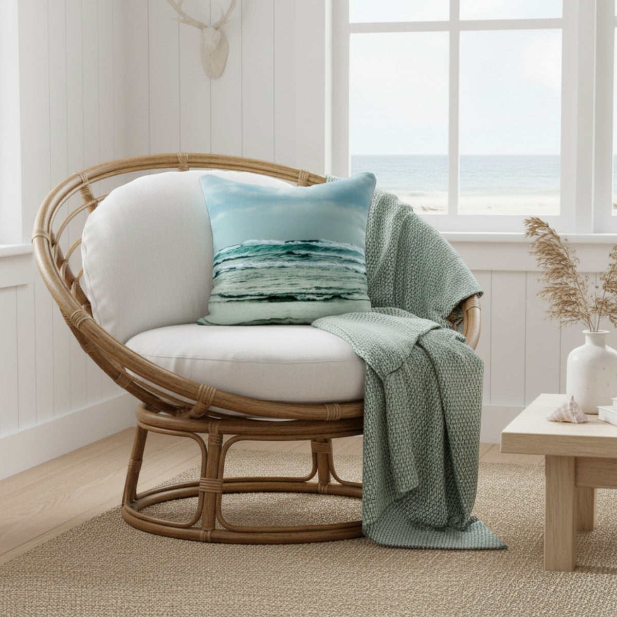 A round woven rattan lounge chair with white cushions, styled with a light blue and teal ocean-patterned throw pillow and a sage green knit throw blanket, positioned in a bright, modern coastal living room next to a window.