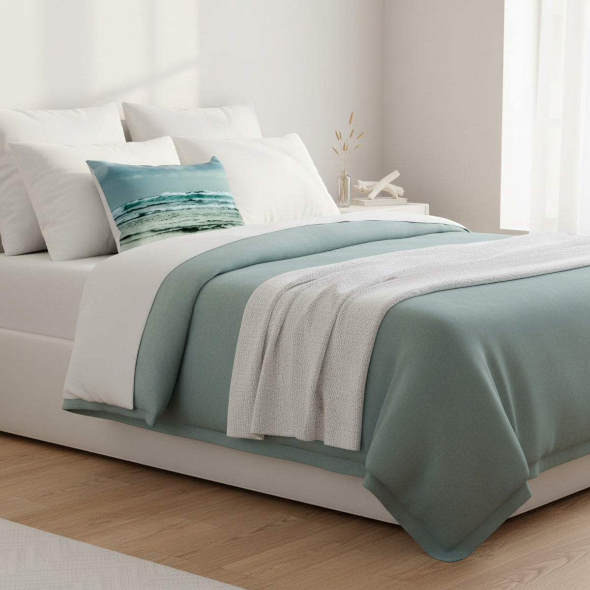 A neatly made bed in a bright bedroom setting, styled with white bedding, a sage green throw blanket, a matching green bed skirt, and a decorative teal and white striped accent pillow.
