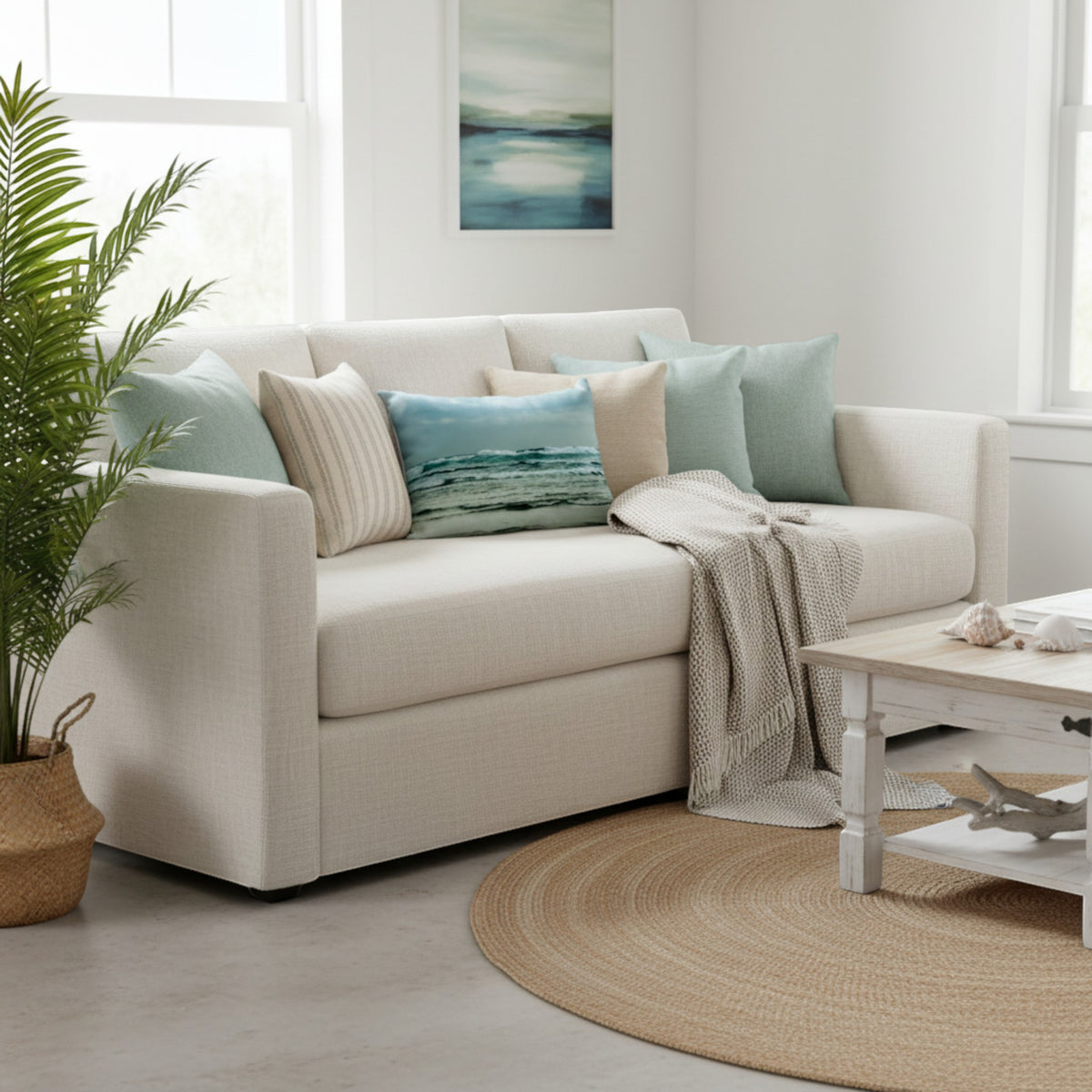 A light beige fabric sofa styled in a bright, modern living room with mint green, blue, and cream throw pillows, a beige knit throw blanket, a potted palm, and a white coffee table on a round jute rug.