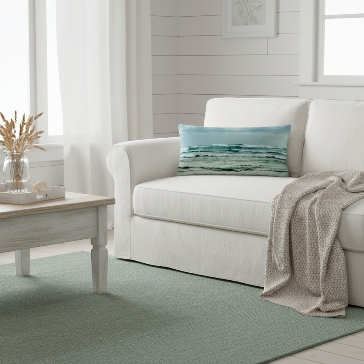 A white slipcovered sofa in a bright living room, styled with a rectangular ocean-print throw pillow and a draped beige knit blanket. A light green rug covers the floor, and a white wooden coffee table with a vase of dried grass sits to the left.