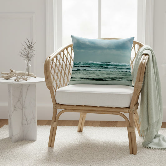 A square throw pillow with an ocean wave print in shades of blue and green, styled on a light wood rattan armchair with a white cushion and a mint green throw blanket in a bright, modern room.