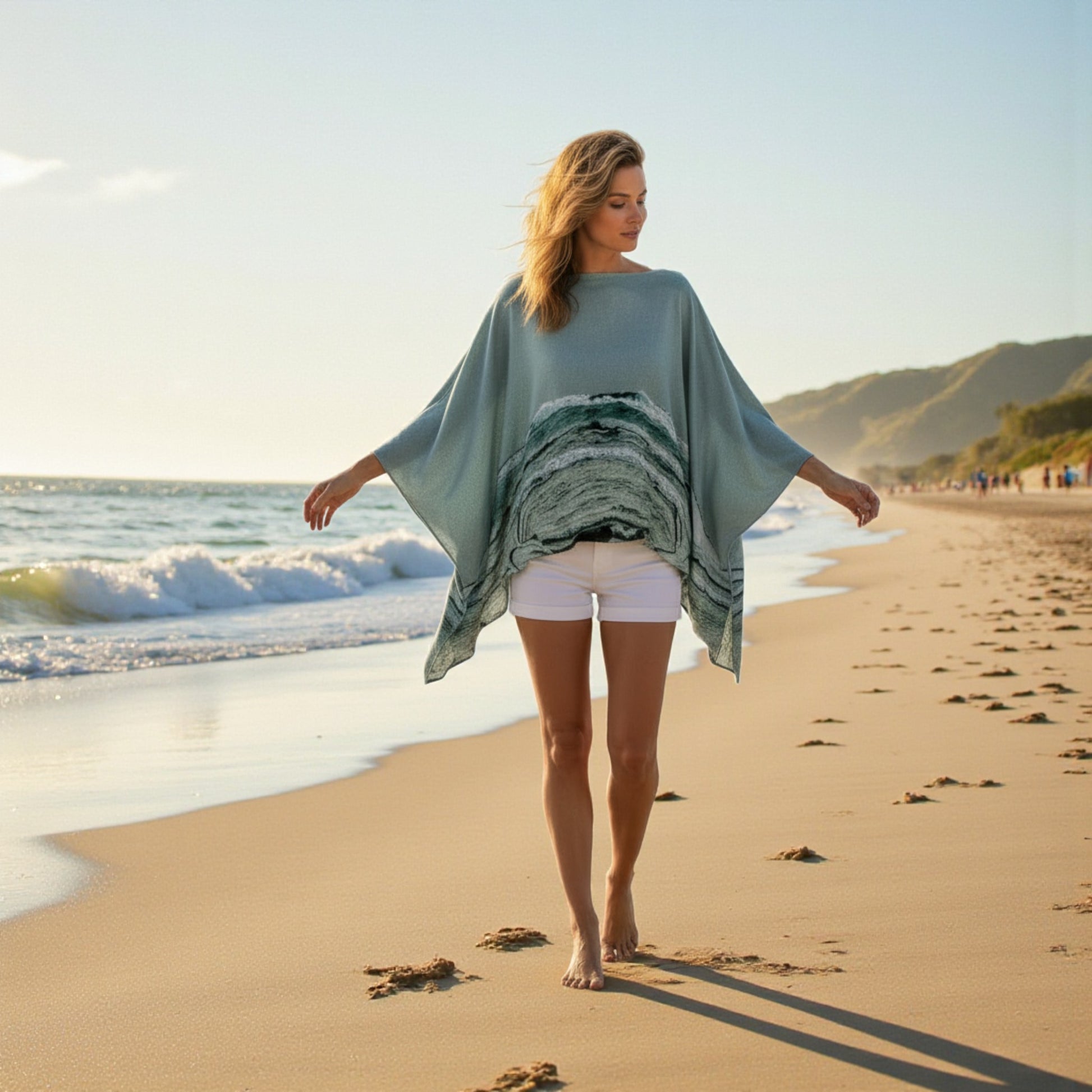 Woman walking on a beach wearing a light blue poncho with ocean patterns.