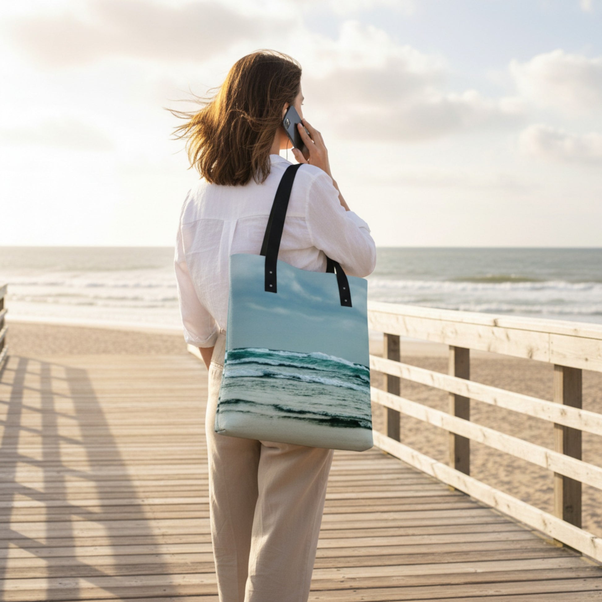 Woman on a wooden boardwalk by the ocean holding a phone to her ear with a beach-themed tote bag.