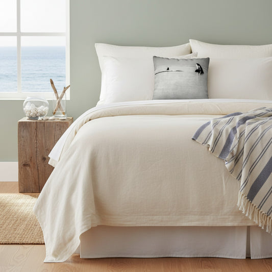 A neatly made bed dressed in off-white bedding, with a striped blue and white throw blanket draped over the corner and a small gray decorative pillow in the center. The room has a pale green wall, a wooden side table with a glass bowl and driftwood, and a window showing the ocean view.