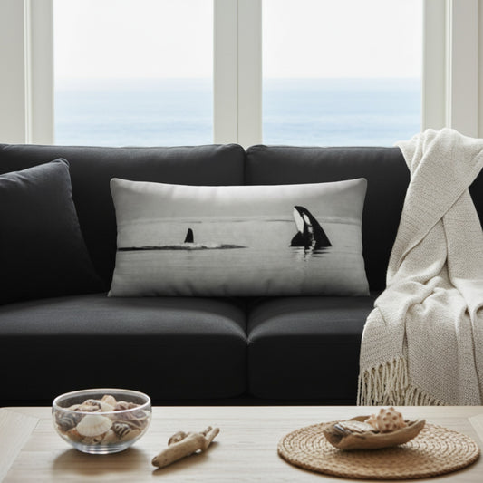 A rectangular throw pillow with a black and white orca print, styled on a dark gray sofa in a living room setting with a window showing the ocean view.