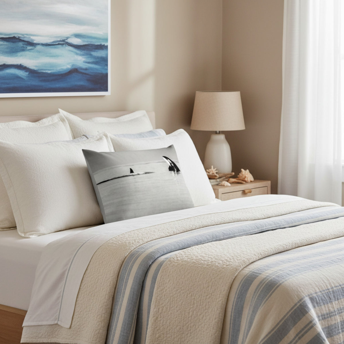 A styled bedroom scene with a bed dressed in white linens, cream bedding, and a blue-and-white striped throw blanket. A square decorative pillow with a beach scene print rests on the bed.