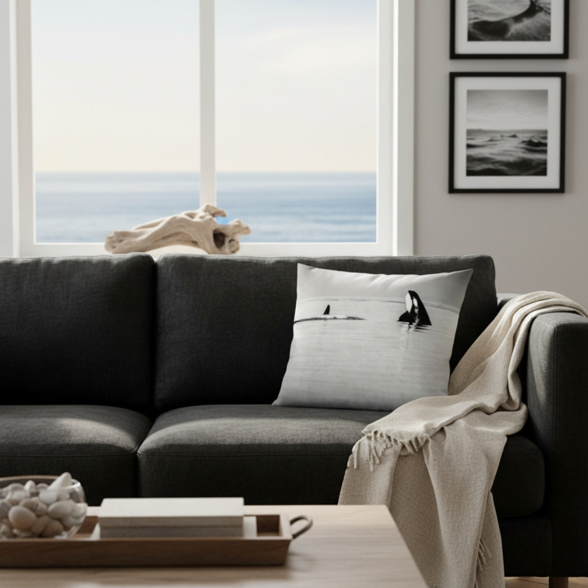 A black fabric sofa in a bright living room with an ocean view, featuring a white throw pillow printed with a minimalist black-and-white penguin and surfboard scene. A beige throw blanket is draped over the arm, and framed wall art hangs on the wall.