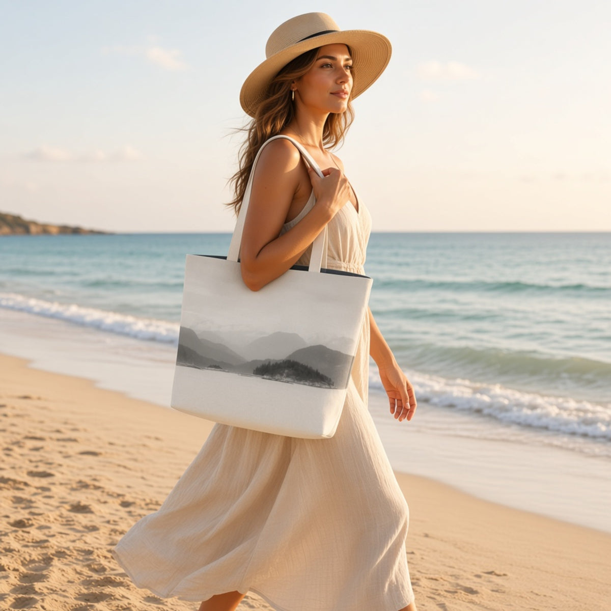 A woman in a cream dress and wide-brim straw hat walks on a sandy beach carrying a large white tote bag with a black and gray mountain landscape print.