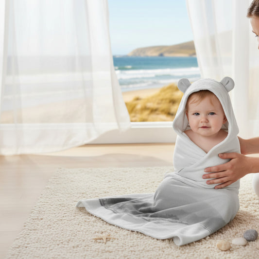 Baby wrapped in a white hooded towel with bear ears, sitting on a carpeted floor with a beach view.