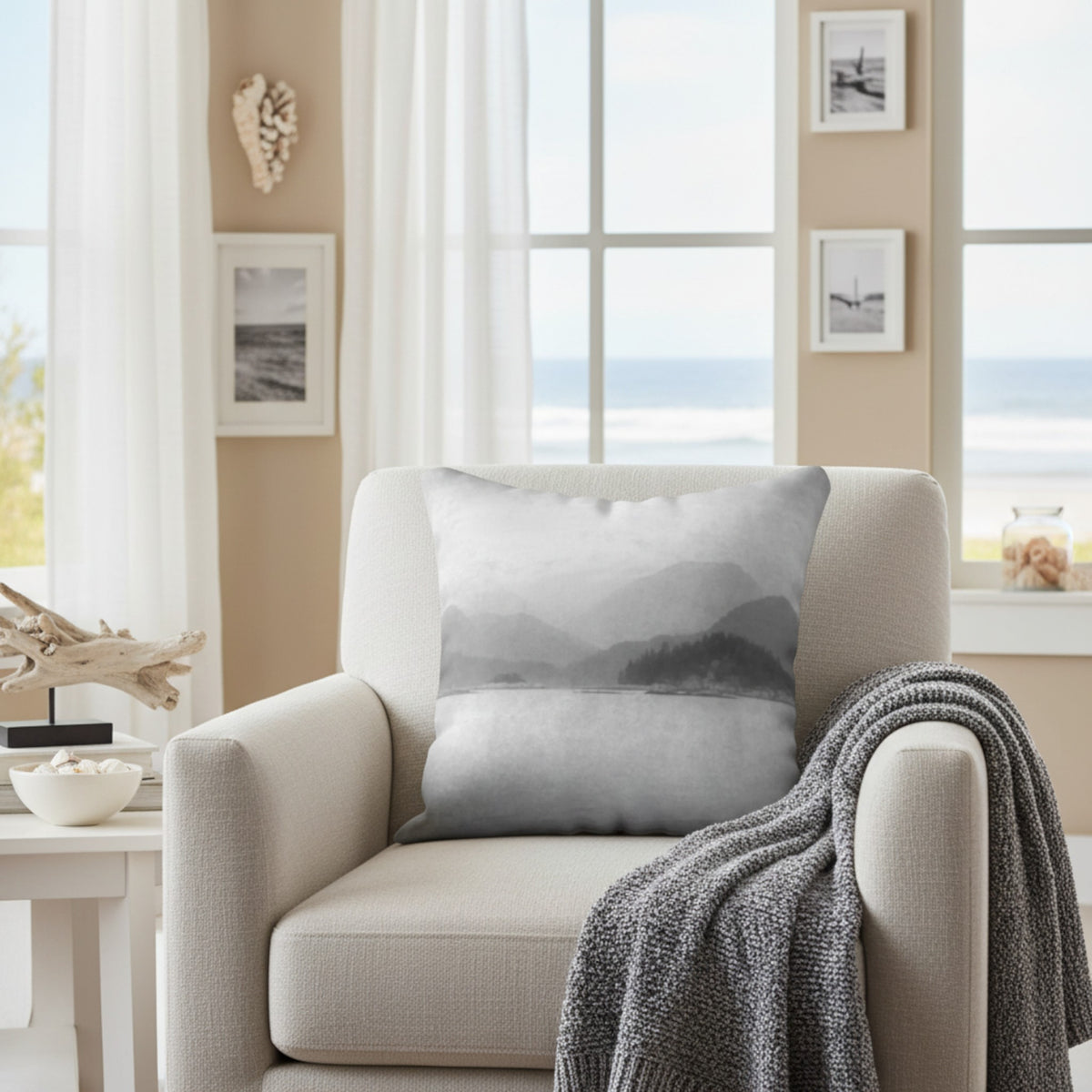 A square throw pillow with a gray and white misty mountain landscape print, styled on a beige armchair in a bright, modern living room with a gray knit blanket draped over the arm.