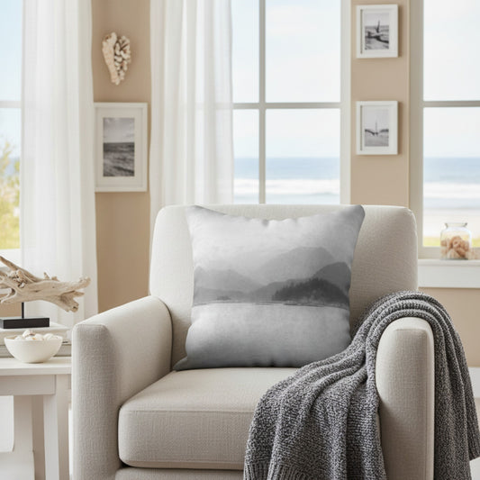 A square throw pillow with a gray and white misty mountain landscape print, styled on a beige armchair in a bright, modern living room with a gray knit blanket draped over the arm.